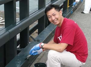 A man in a red t-shirt using a paintbrush on railings along a pier.