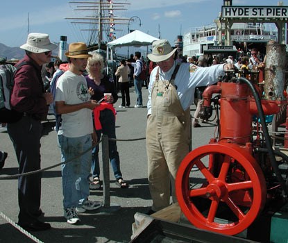 Park volunteer on Hyde Street Pier standing next to a red Hick's engine and talking to interested park visitors.