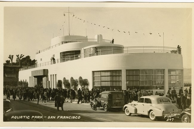 Black and white postcard showing hundreds of people and parked cars outside the entrance of the Aquatic Park Bathhouse in 1939.