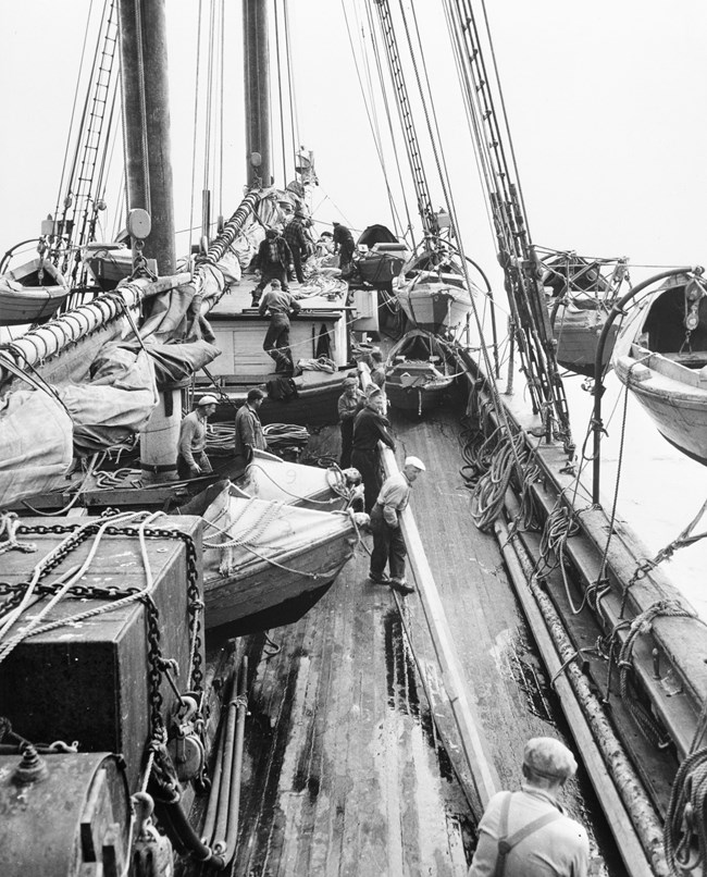 historic image of men on the deck of a ship loading smaller fishing boats into the water
