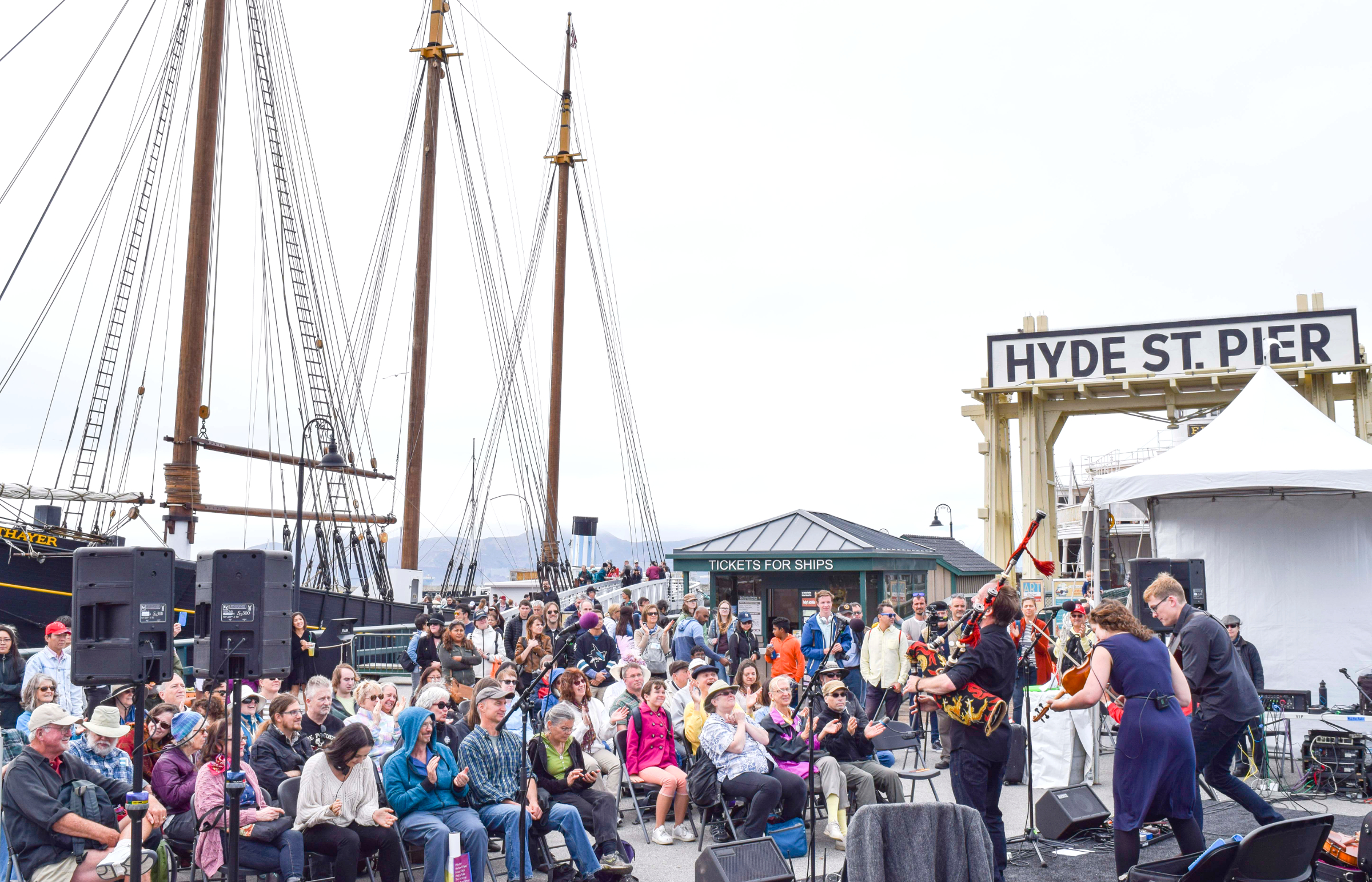 Crowd of people sitting in folding chairs and standing to watch a band perform on a stage near the Eureka ferryboat gangway.