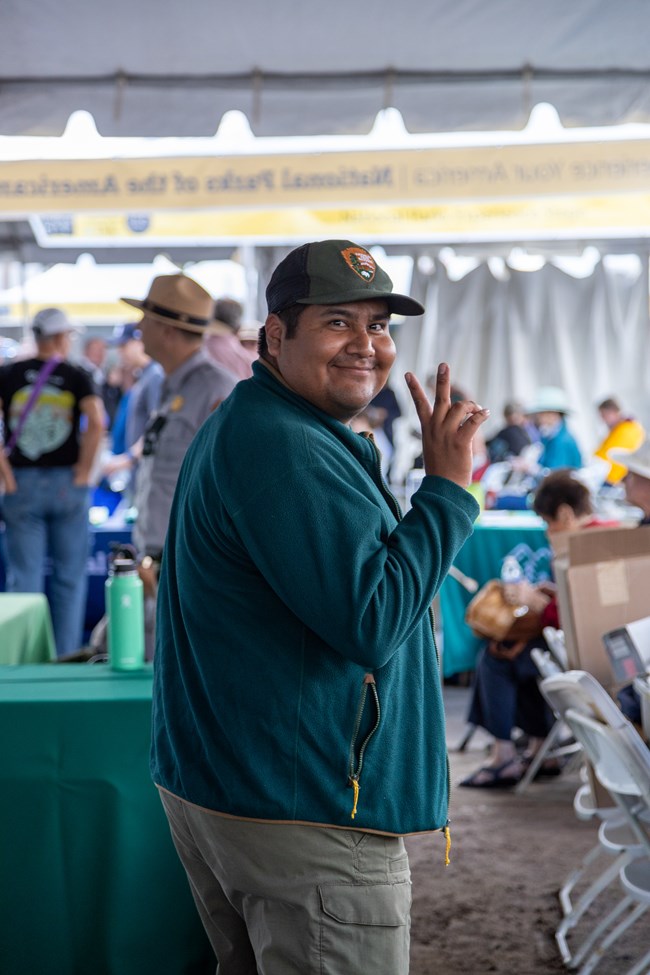 Vertical Image. Front a center a young adult man turns over his right shoulder to look back at the camera. He has a large smile and is holding up a peace sign as he walks away. He is wearing a green NPS hat, green felt jacket, and dusty green pants.