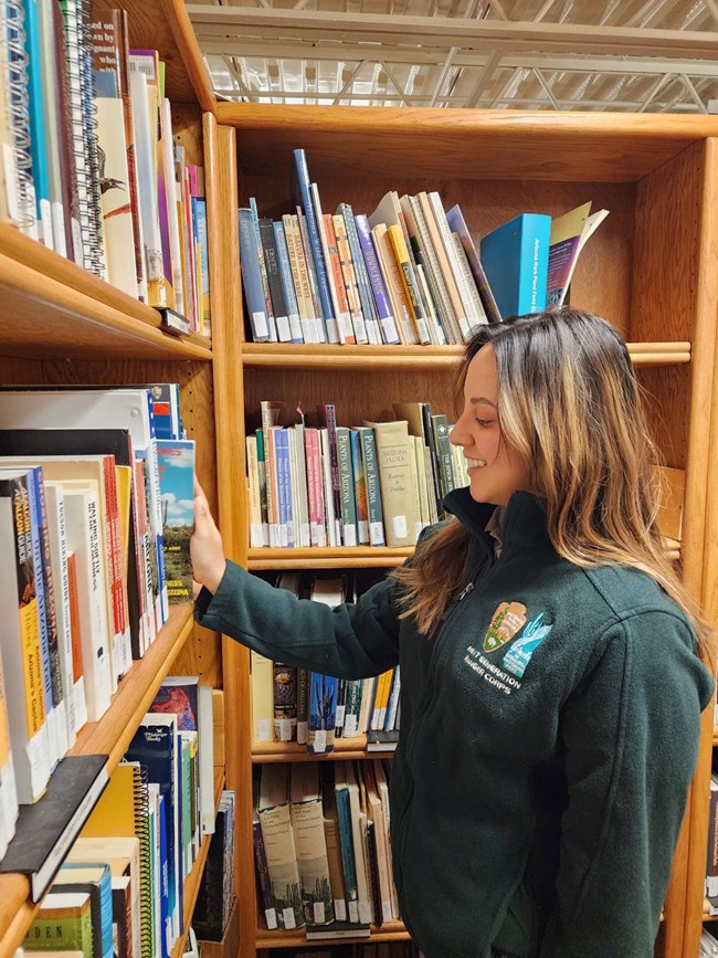 Vertical image. In the lower right quarter of the image there is a young female intern, wearing a green jacket with various logos on the chest. She is reaching out and pulling a book from a tall wooden bookshelf that surrounds her on all sides.