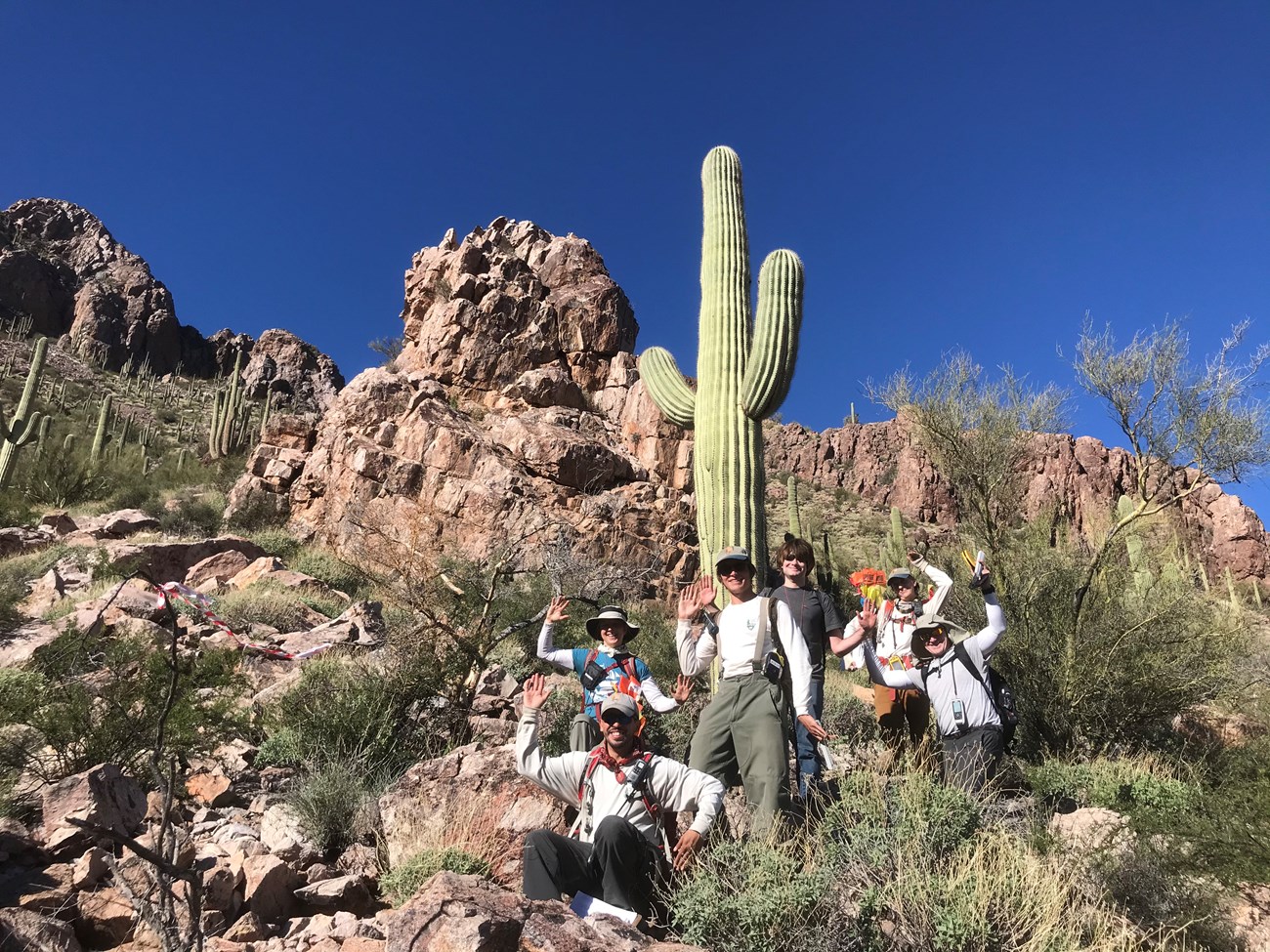 A group photo where everyone is posing like a saguaro. Behind them is a tall saguaro and a nice mountainous view.