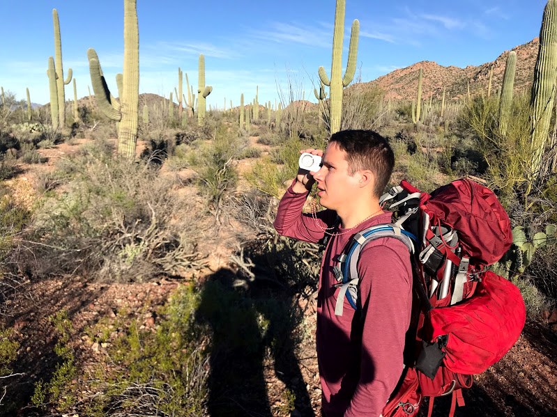 A man with his backpack looking through a clinometer. On the background are saguaros and lots of vegetation.