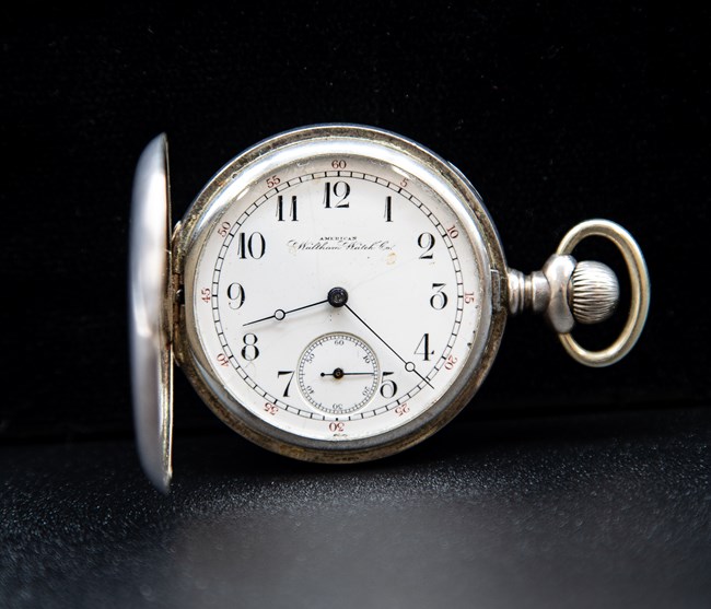 A silver pocket watch, with the name American Waltham Watch Co. written on the face, and the hour hand pointing to just above the 8, and the minute hand pointing between 4 and 5.