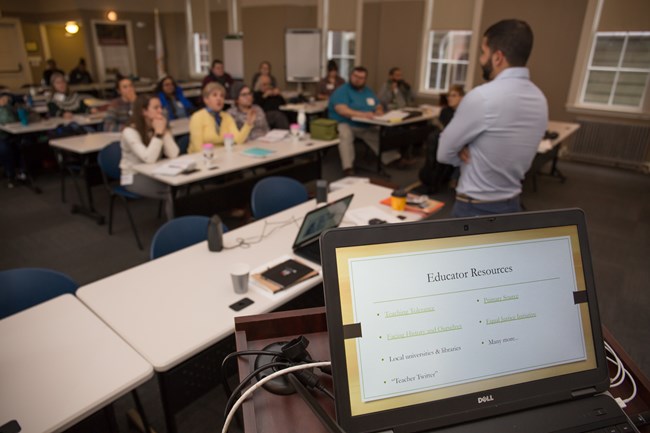 Open laptop computer with screen reading 'Educators Resources' in foreground. Out of focus in background is a man with his back to the camera, standing at the front of a room filled with people sitting at tables.
