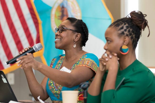 lt= Upper profiles of two women. Woman to the left smiles and speaks into a microphone.