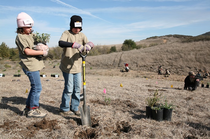 A kid digs a hole with a shovel and another kid behind holds a plant