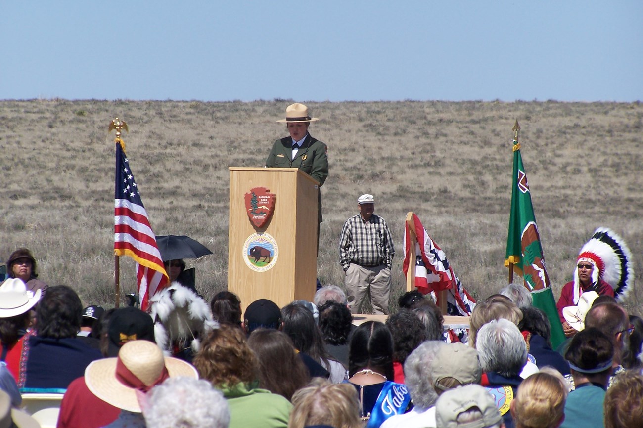 A uniformed leader speaks at a podium in front of a prairie landscape
