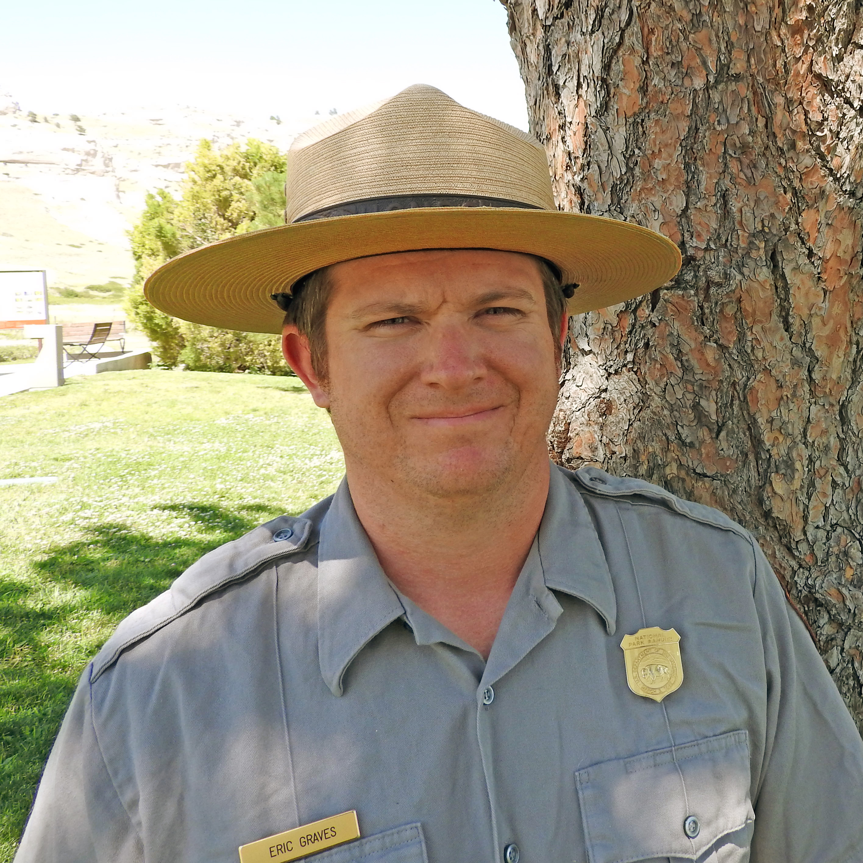 A park ranger stands in the shade of a pine tree.