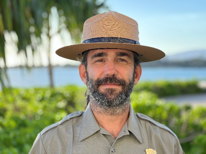 A bearded man is seen from the chest up. He wears a park ranger hat. Water and vegetation are out of focus in the background.