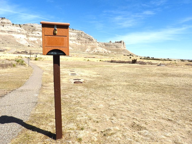 A brown dog waste bag dispener has black dog waste bags protruding from its bottom.