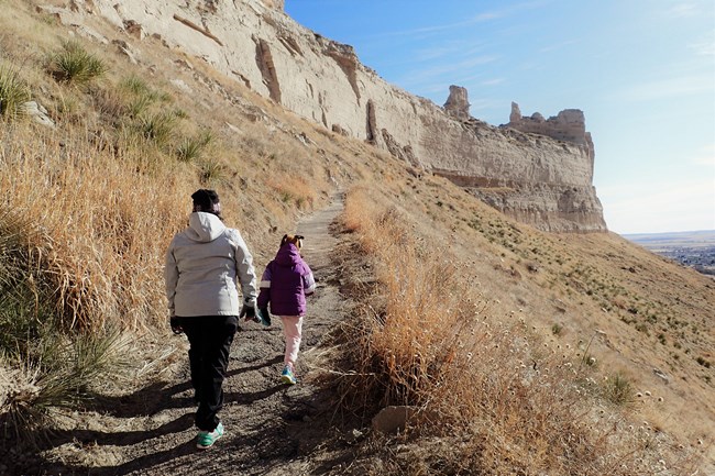 A pair of hikers ascends a paved trail towards a tunnel carved into a sandstone bluff.