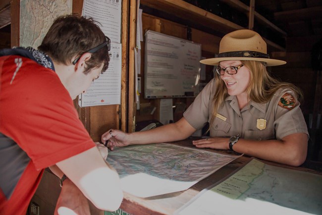 A female ranger leans stands at a desk, pointing out trails on a map to a visitor.