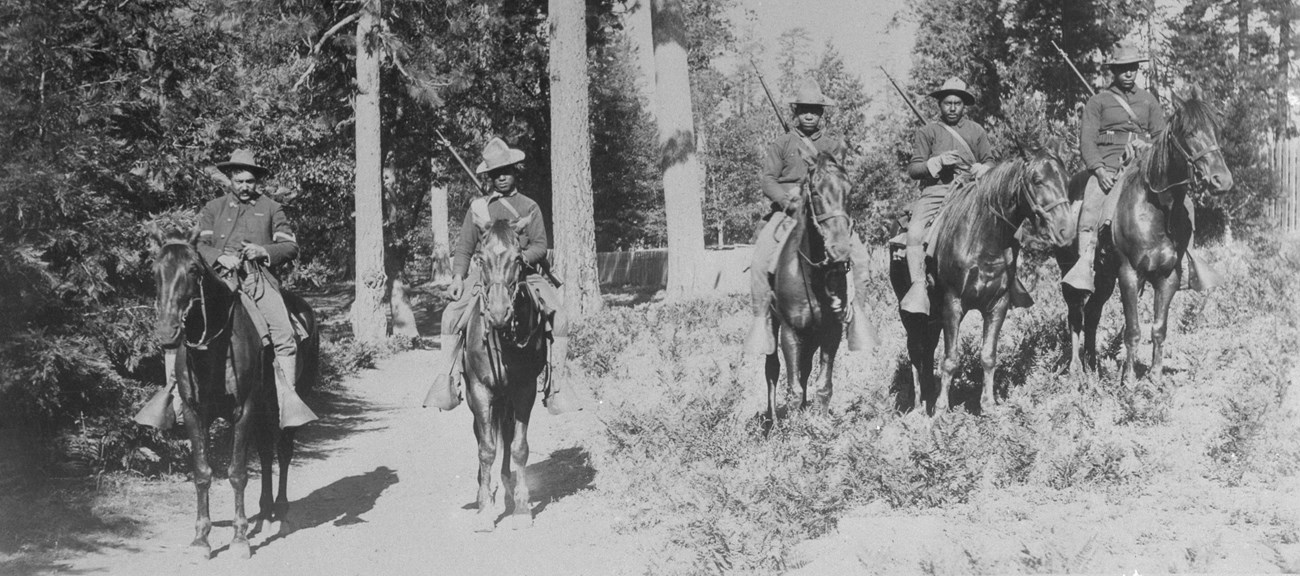 A troop of Buffalo Soldiers mounted on horseback in a forested area.