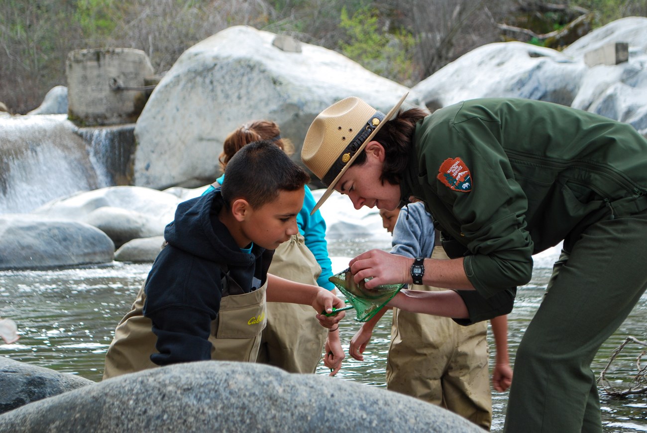 Next to a river, a park ranger inspects the contents of a young student's fishing net.