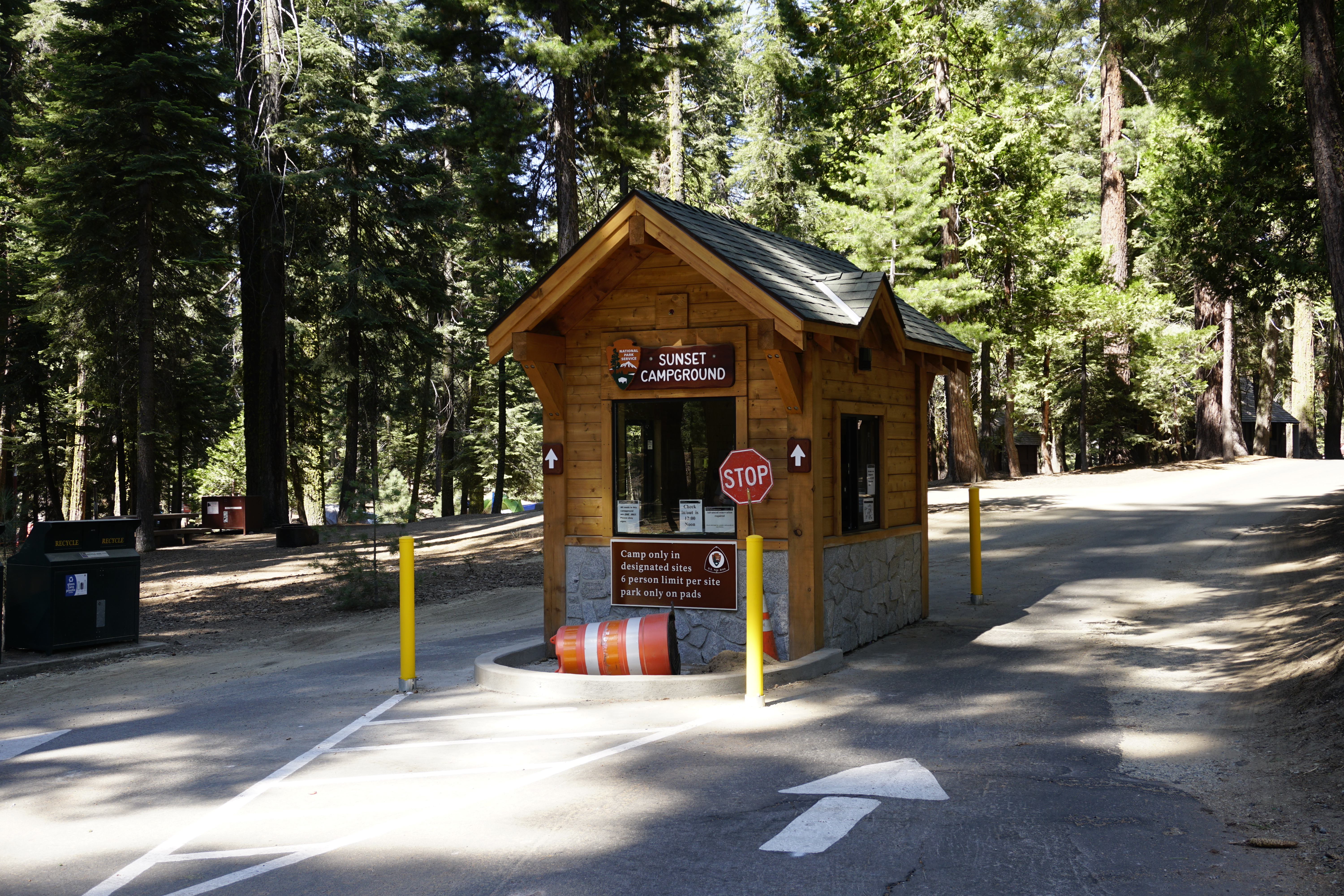 A small wooden building sits into he middle of a grey road. Behind are campsites and large green trees.