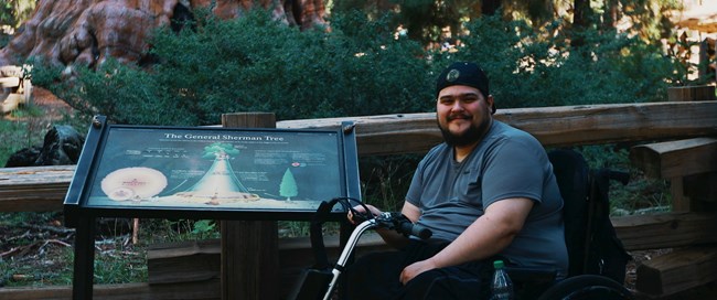 A man in a wheelchair stands in front of an accessible wayside sign.