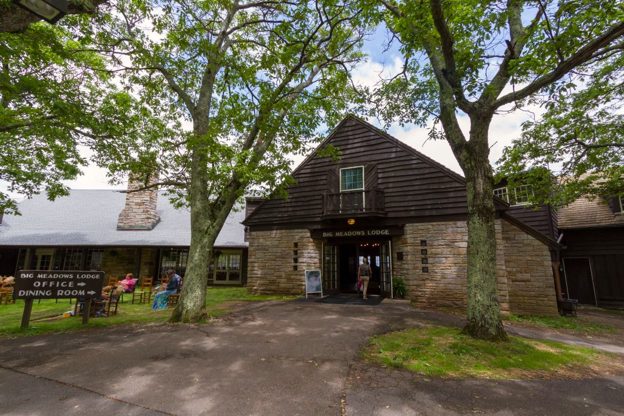 A brown stone and dark brown sided building labeled "Big Meadows Lodge" stands in the center. A woman walks through a doorway in the center of the building. A window above the doorway. In front of the building are two large trees and green grass on either