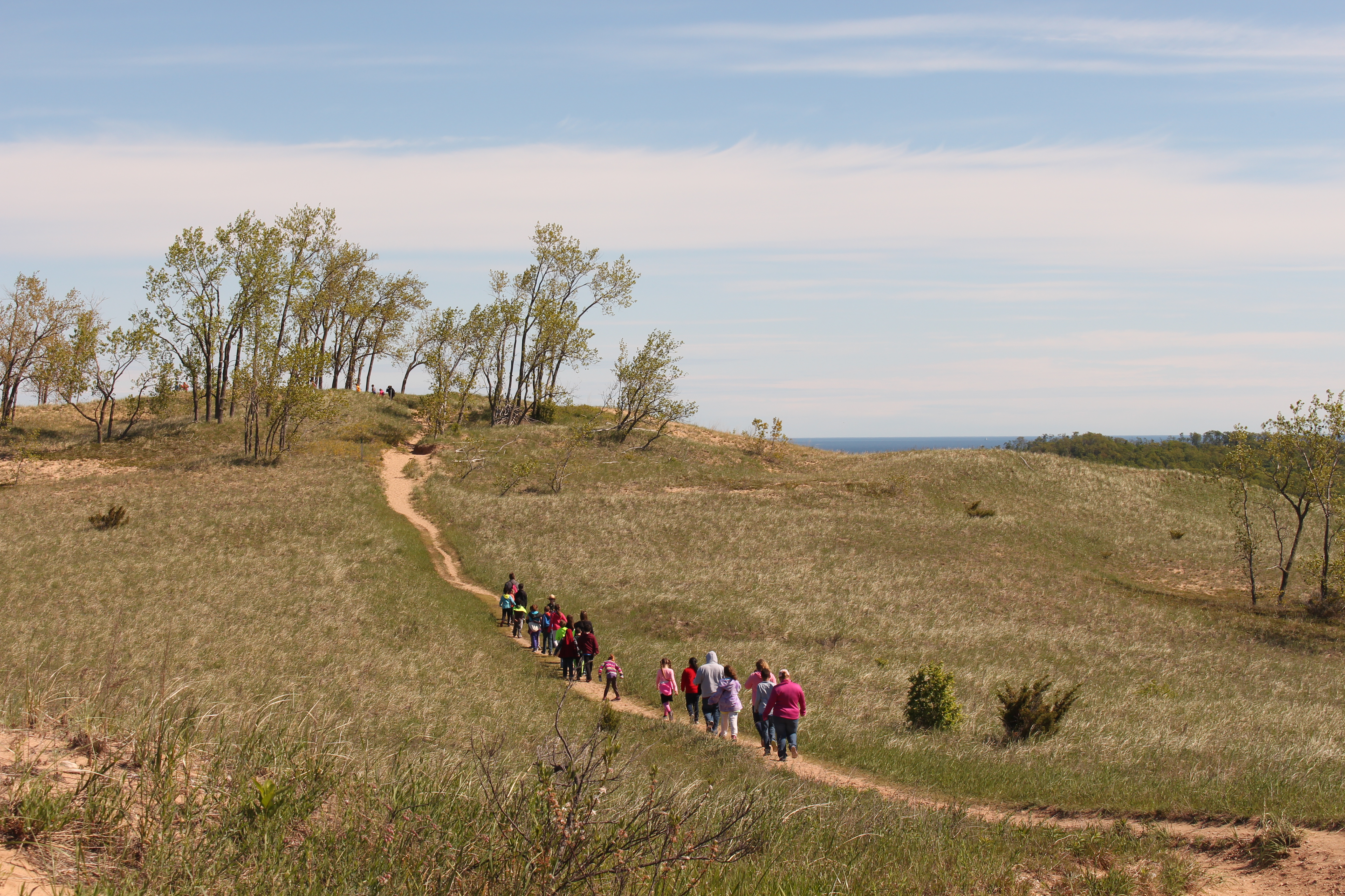 A group of students hikes on a sandy trail through grassy dunes. A small grove of trees and a lake is visible in the background.