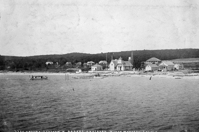 East Side Village (MIA)Tilt North Manitou Island Life Saving Station sits along the beach, including a boathouse and crew quarters, behind the station sits a row of cottages. All the buildings are painted white and stand out sharply from the dark forest in the background.