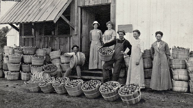 FrankFarm A family of farmers stands in front of a large wooden building, surrounded by baskets full of apples.