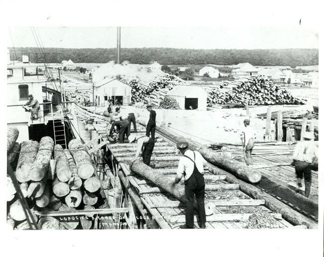 NMI Mill Operation Workers move along a long dock loading cut logs onto a ship. In the background are many buildings and tall stacks of logs. Words along the bottom read: “Loading cargo of logs at the Manitou.”