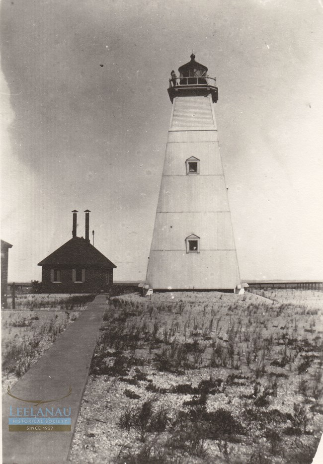 nmi lighthouse A tall, pillar shaped lighthouse stands on a sandy beach. A white tower with a black top. Behind it sits a shorter fog signal building with two smokestacks protruding from the roof. A vigilant lighthouse keeper stands on the catwalk at the top.