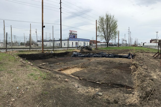 A square pit in the ground. Electrical poles and businesses lie in the distance behind.