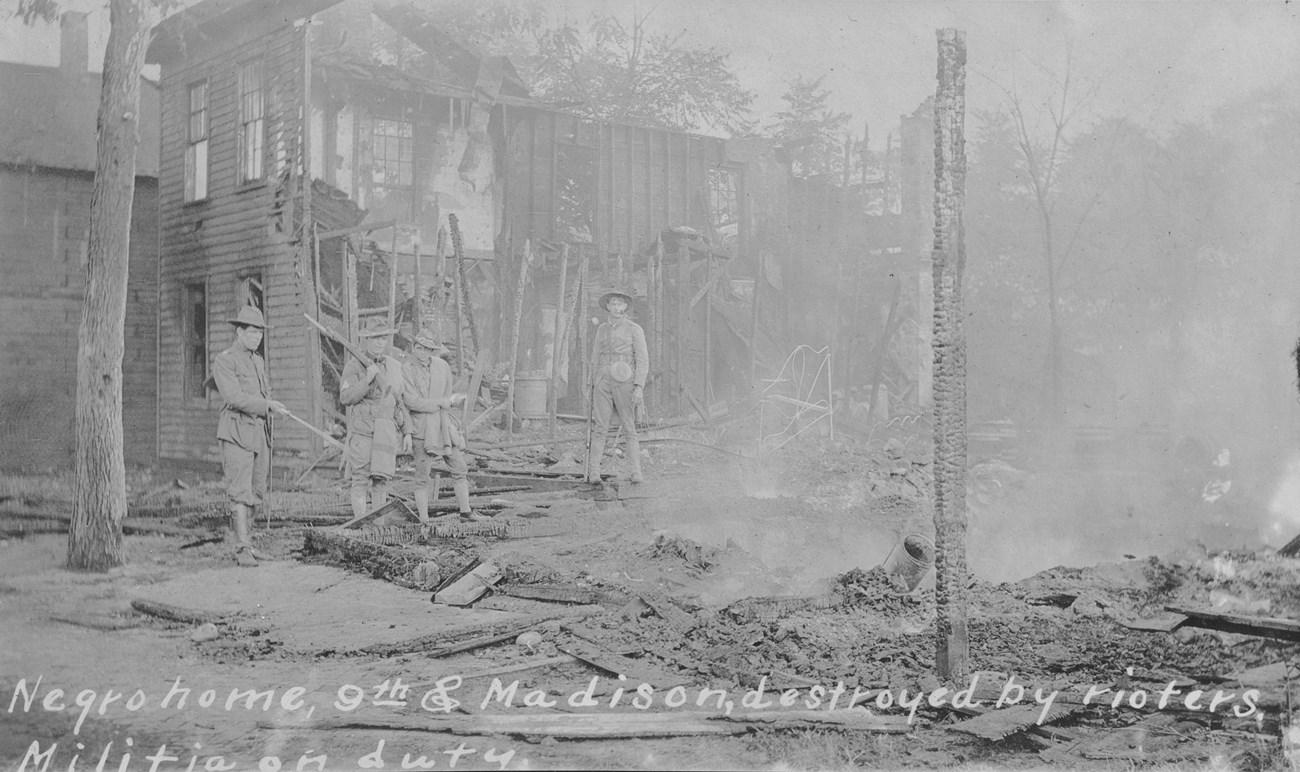 Historic black and white photograph with text that reads, “Negro home, 9th & Madison, destroyed by rioters, militia on duty.”