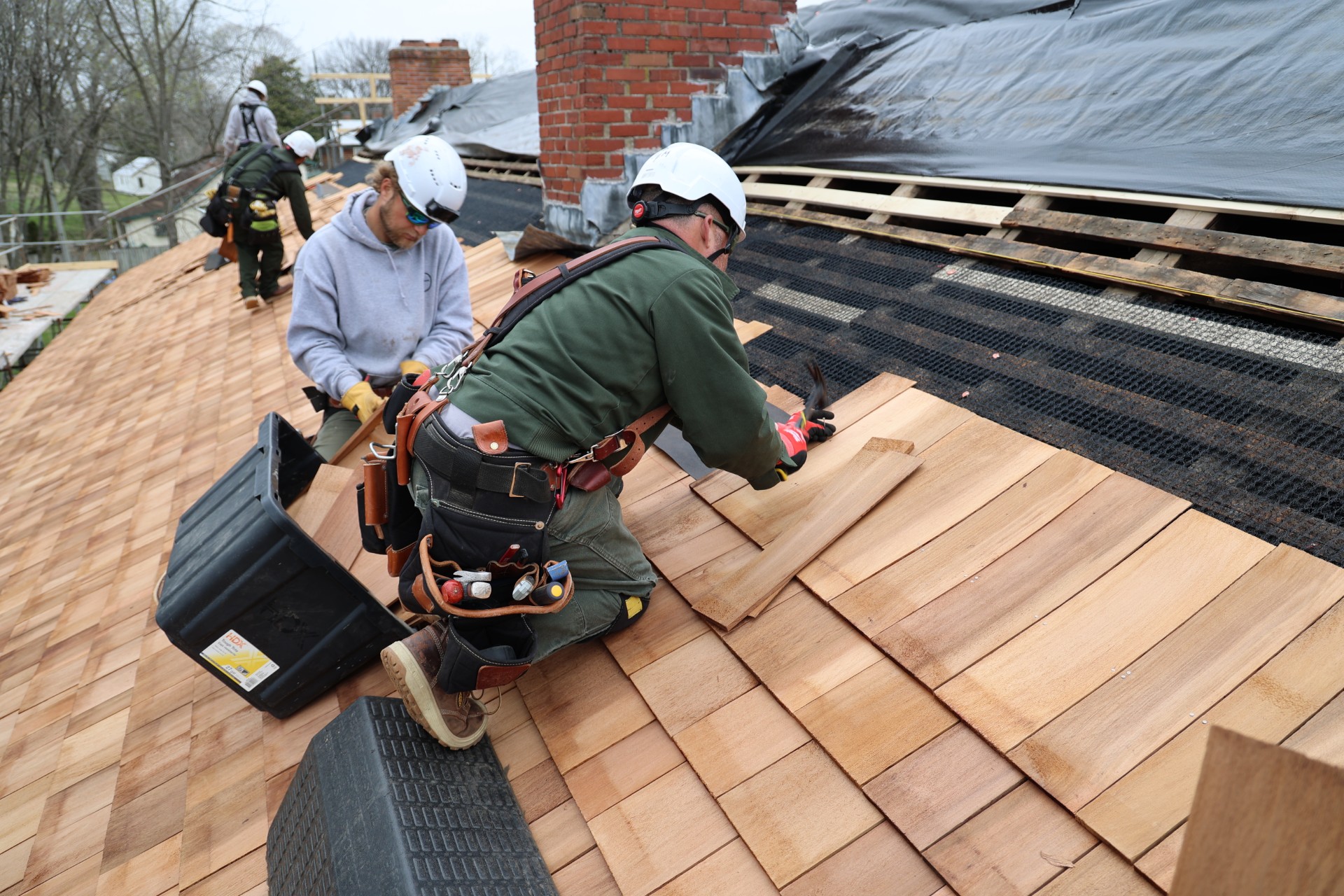 Three individuals in hard hats and gloves replacing wooden shingles on a roof.