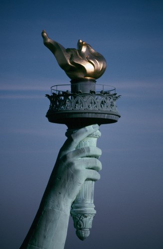 Outstreched right hand holding a torch topped by a 24k gold leaf covered flame