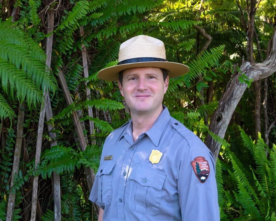 Ben Hayes A man wears a National Park Service gray shirt and flat hat in front of a stand of ferns.