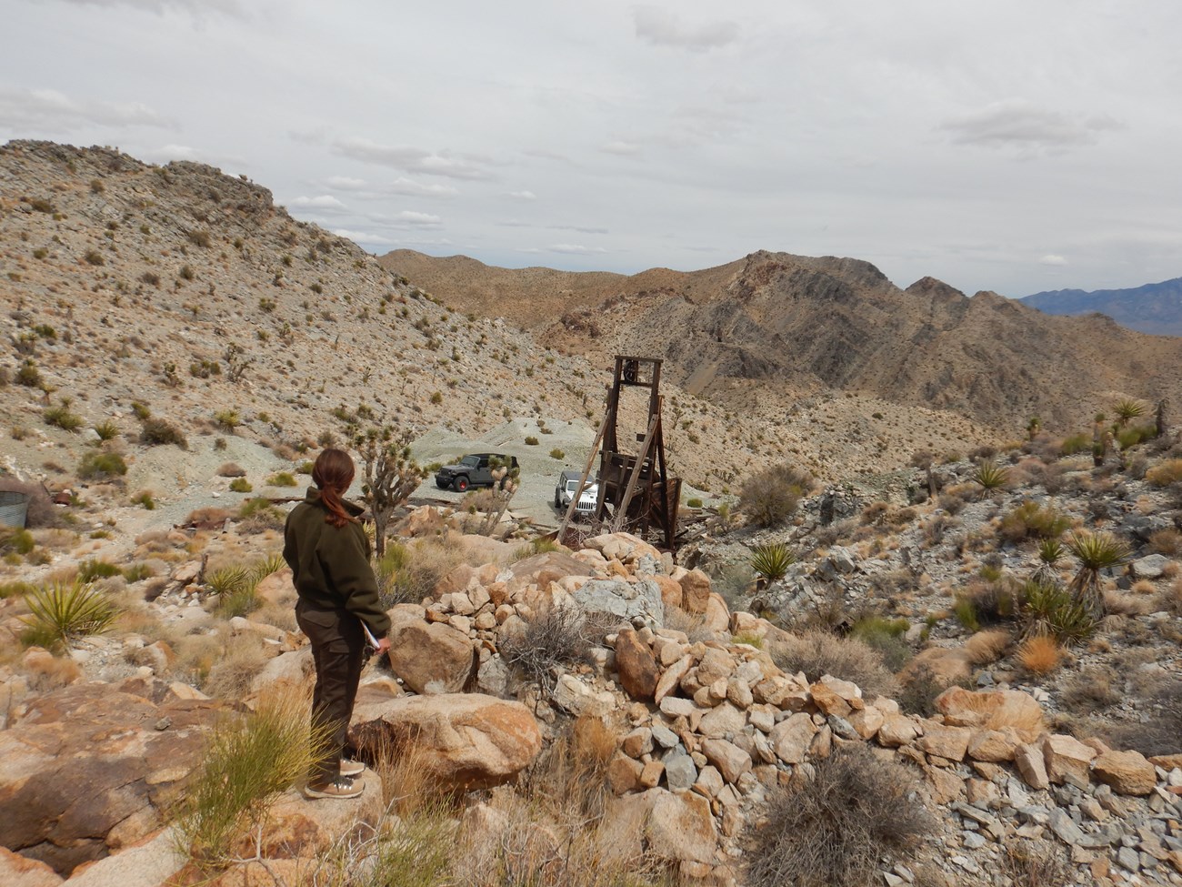 photo of a park employee inspecting a mine site in a rocky desert landscape. In the distance, 2 Jeeps can be seen parked near a mining headframe.