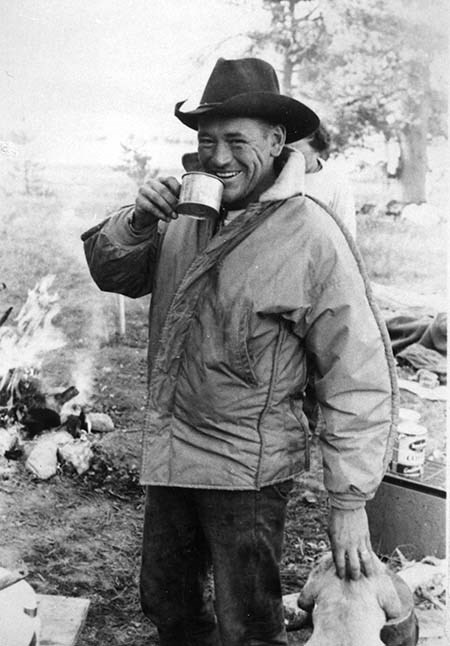 A man in a cowboy hat and jacket smiles as he drinks from a tin cup near a fire.