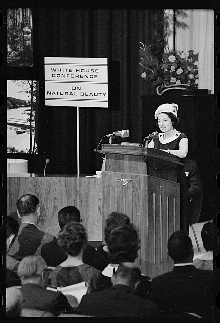 Lady Bird Johnson at Conference for Beautification A seated audience watches a woman who speaks from behind a tall podium with two microphones, next to a sign that says "White House Conference on Natural Beauty"