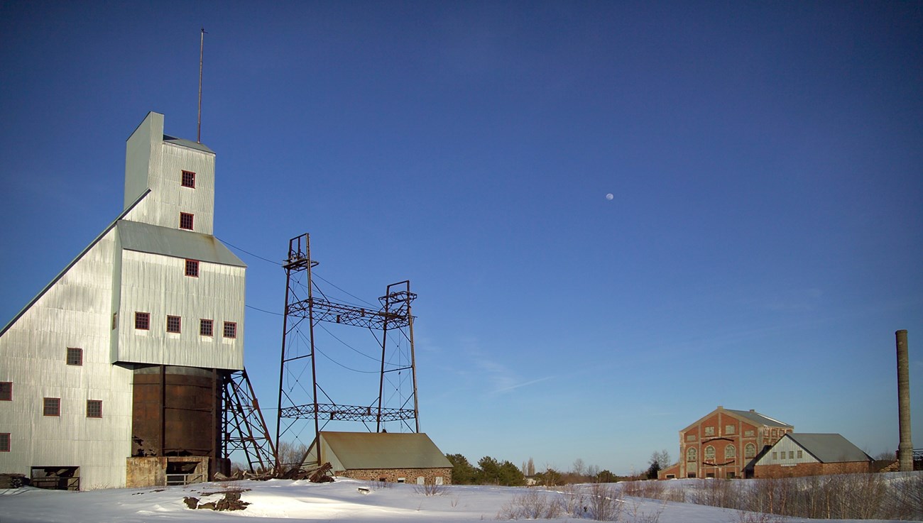 Snow covers the site of the former Quincy Copper Mine The No. 2 shaft-rockhouse , metal and wire of the pulley stands, and large brick hoist house with smoke stack are spaced in a level area