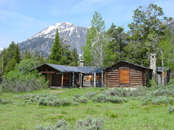 Single-story log cabin with boarded windows is surrounded by scrub, trees, and snow-capped mountains.