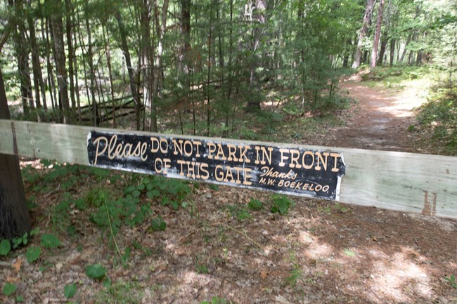 A sign on a wooden gate across a trail in the woods says "Please do not park in front of this gate. Thanks, M.W. Boekeloo"