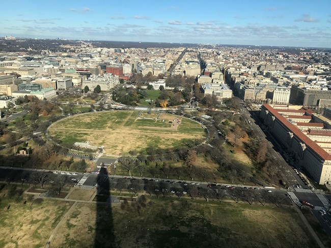 Aerial view of the Ellipse, a circular area of turf, and surrounding park and city. The narrow shadow of the Washington Monument is in the foreground and the White House is in the center.