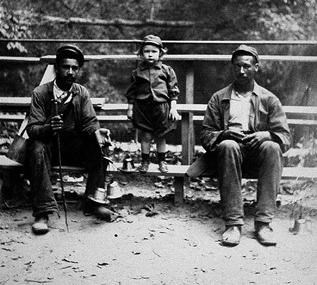 Two African American men are seated on a wooden railing along a path. A child stands between them.