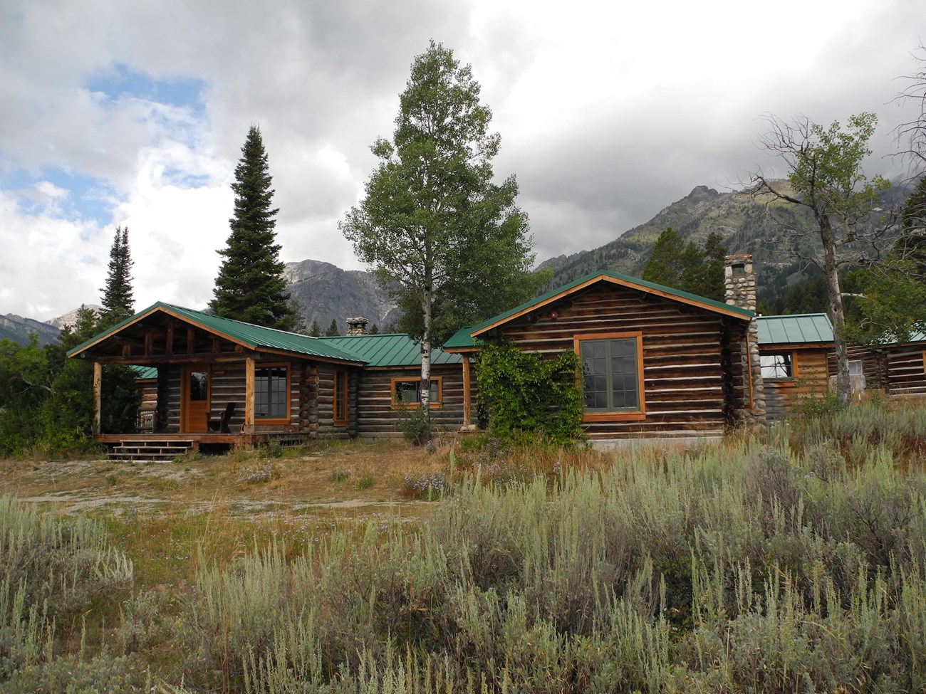 Sagebrush grows in front of a one-story log cabin, surrounded by several trees and tall mountains.
