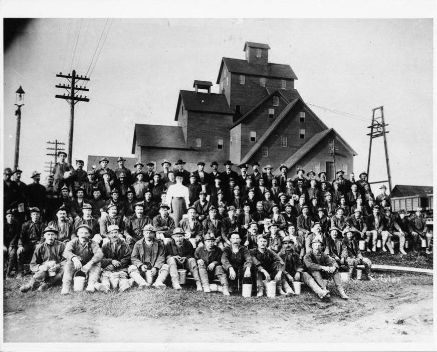 A large group of workers pose in five rows, seated and standing, in front of a tall industrial shafthouse.