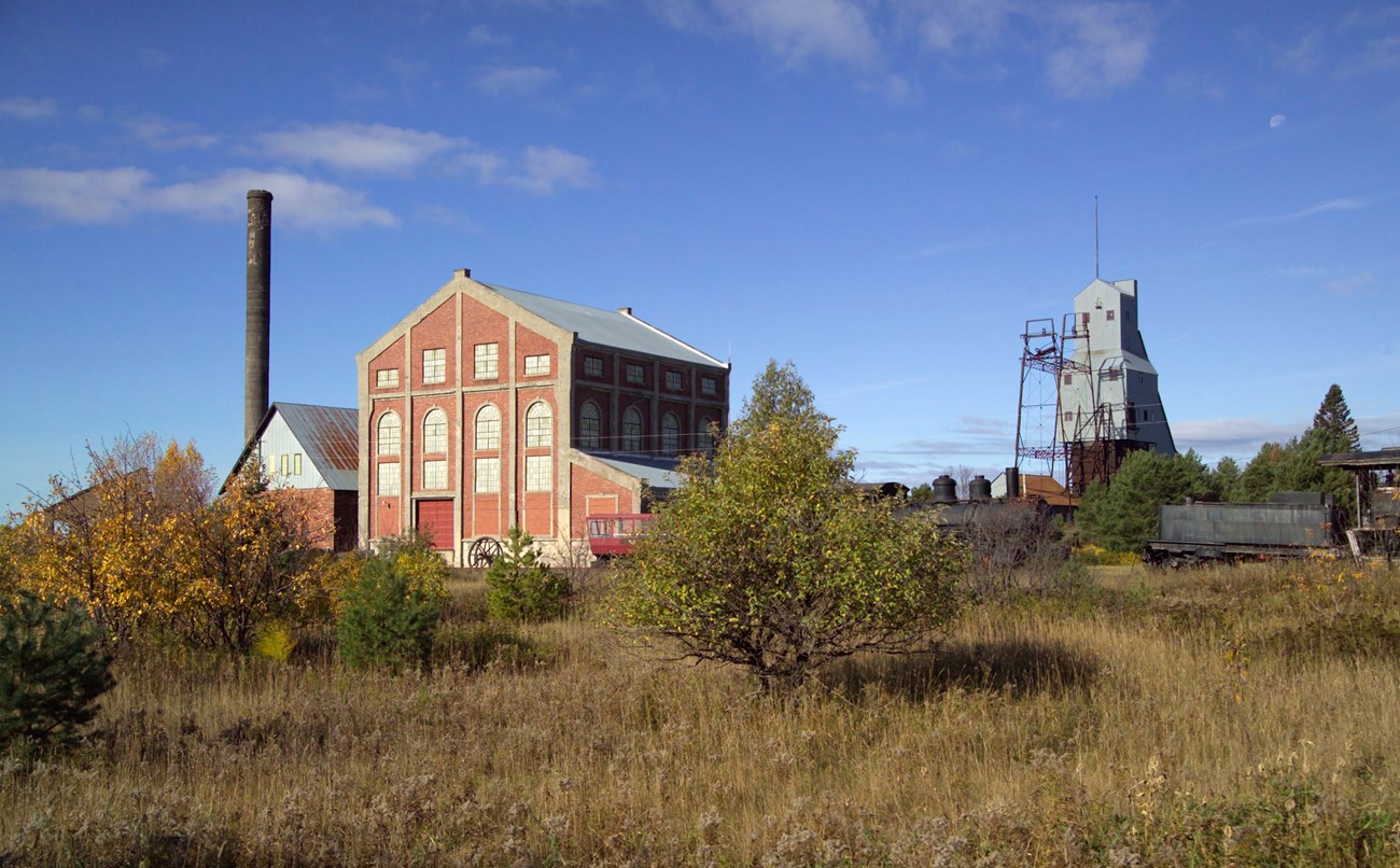 Tall industrial structures, including a brick building, tower, and hoist, surrounded by a landscape of low trees and grass in fall colors