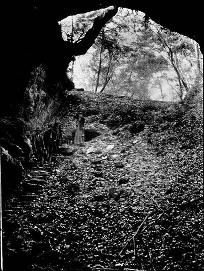 Black and white image of a person standing on a staircase at the entrance of a cave, with a wide opening looking toward trees and sky.