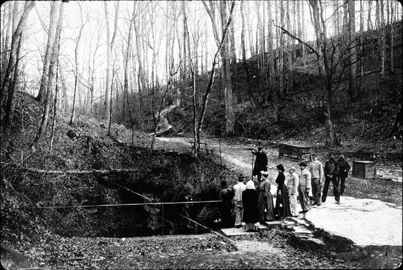 Black and white photo of a group of people standing at a railing around a hole in the ground in a wooded area.