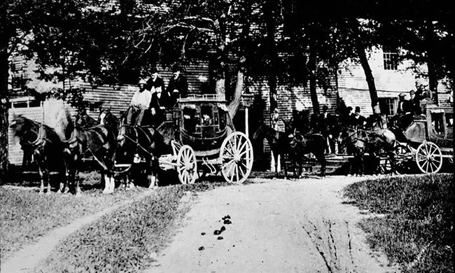 Two horse-drawn stagecoach cars with drivers and passengers stand along the side of a dirt road.