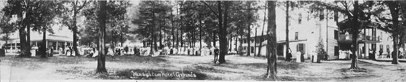 Black and white panorama of Mammoth Cave Hotel Grounds with people posed in the lawn, under scattered trees, and on the porch of the two-story hotel