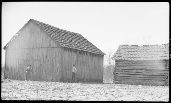 Black and white photo of two men stand beside a wooden barn and log pen